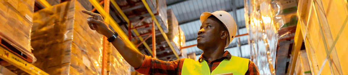 In a warehouse, a man wearing a white hard hat, yellow safety vest, red flannel shirt and blue jeans looks for a product while holding a tablet. In the background two warehouse workers wearing yellow hard hats and safety vests are having a discussion.