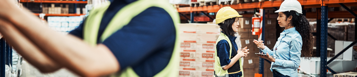 Distribution warehouse workers, a man on the left using his hands and women on the right talking. Both workers wear blue shirts and dark pants.