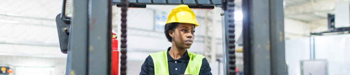 A woman wearing a reflective safety vest, dark polo shirt and hard hat steers a forklift.