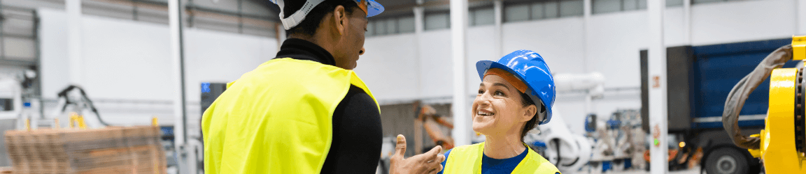 Two workers in yellow vests and blue hard hats talk to eachother on a production room floor