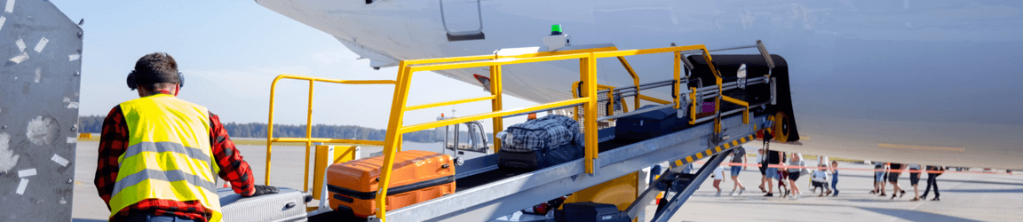 An airport worker wearing a yellow vest loads bags onto an airplane outside.