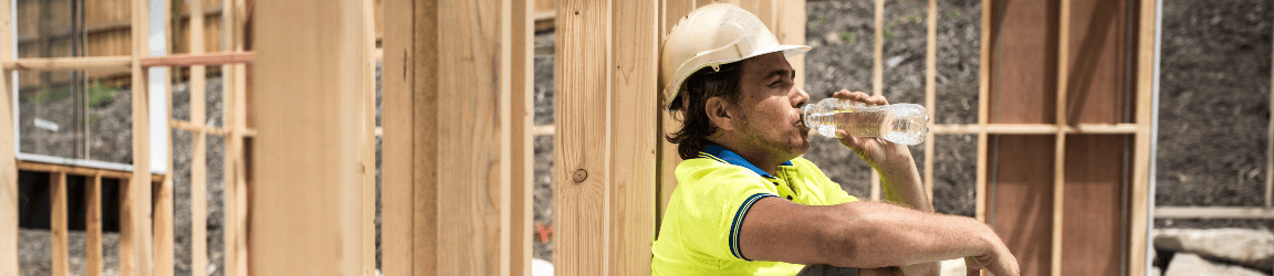 A male construction worker wearing a hard hat sits next to his tool bag while drinking water on a hot day.