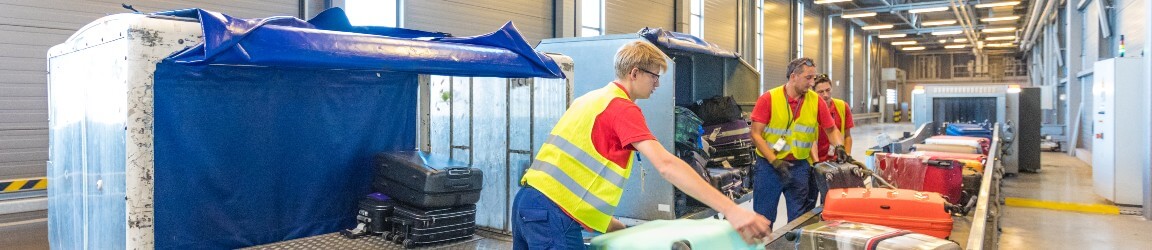 Inside a warehouse, two men facing towards the camera wear orange safety vests and grey jeans. They speak to a woman wearing a yellow safety vest while she holds a tablet.