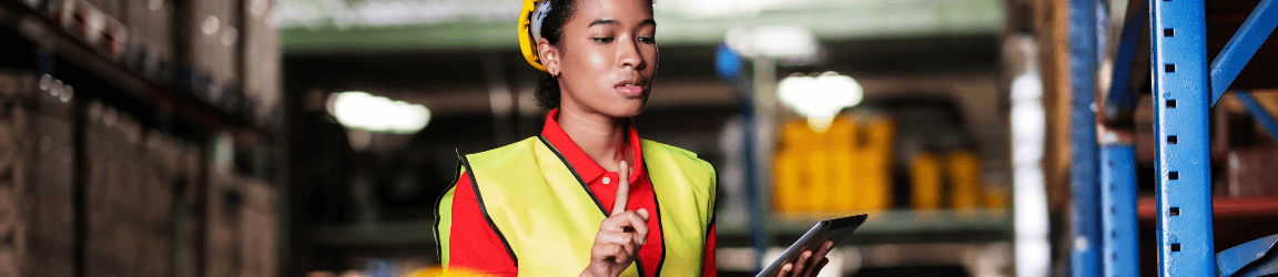 In a warehouse a man and woman wear hard hats and safety vests while logging inventory data into tablets.