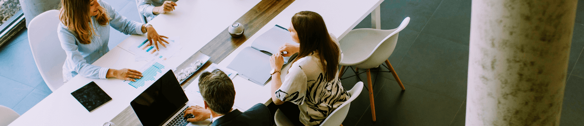 An overhead view of a business meeting shows 5 people having a discussion at a long white table.