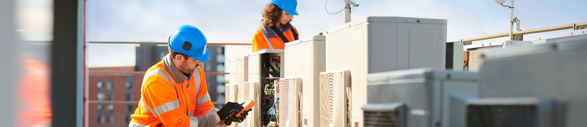Two technicians wearing orange vests and blue hard hats examine heating and air systems on the rooftop of a building.