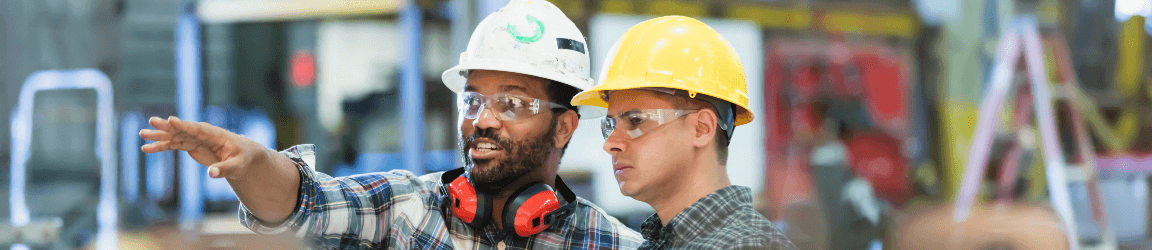 Two factory workers wearing hard hats and safety glasses review blueprints as one worker points out machinery in the distance