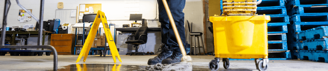In a manufacturing facility, a man — wearing safety glasses, protective gloves, a grey shirt and dark pants — uses a mop and bucket to clean up a liquid spill. He has placed a warning sign near the spill.