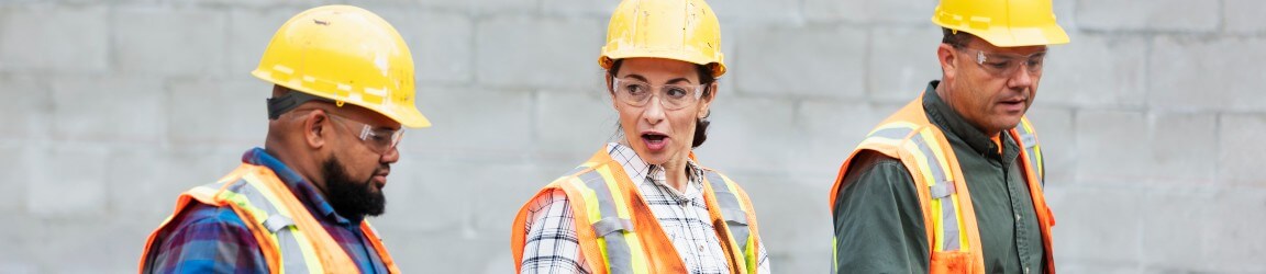On a construction site, a woman wearing a yellow hard hat, safety glasses, plaid shirt and orange safety vests, walks between two men wearing similar attire while talking.